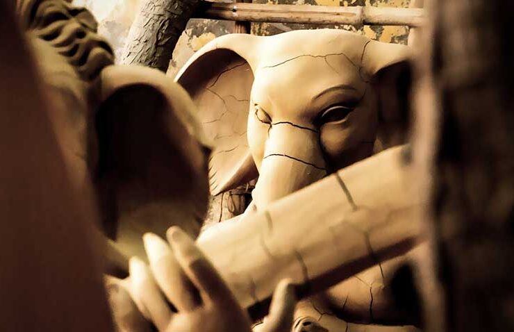 The raw beauty of creation. An intimate, close-up look at a large, unpainted clay Ganesha idol still under construction in a workshop. The prominent cracks across the elephant-headed deity's face, large ears, and trunk emphasize the delicate, labor-intensive process of molding earth into divine art. This raw form, held together with bamboo sticks and ropes, reveals the artistry behind the finished product.