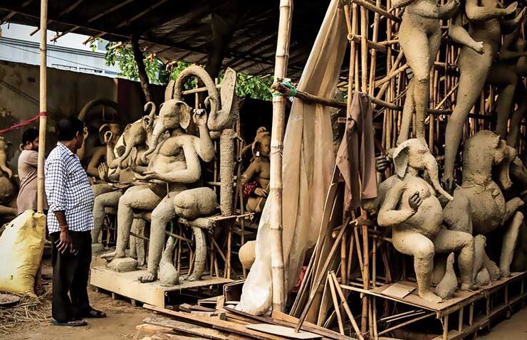 A man stands observing a workshop filled with tall bamboo scaffolding and unpainted clay idols of Lord Ganesha in various stages of creation, highlighting the monumental effort and preparation ahead of the festive season.